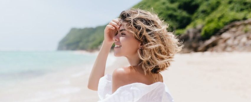 mulher em praia a olhar de perfil para o mar com um cabelo loiro ondulado saudável e hidratado